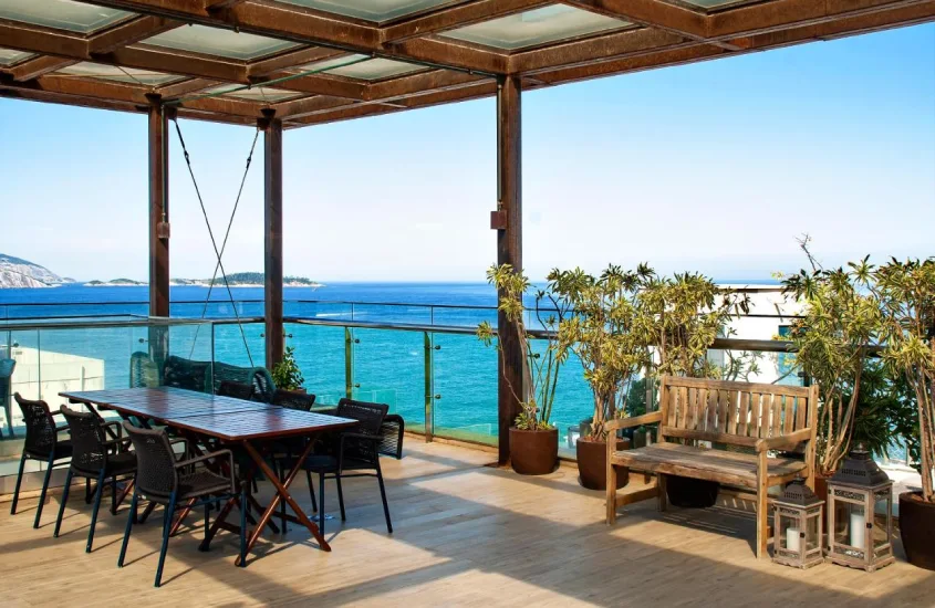 Em um dia ensolarado, cobertura no Arena Ipanema Hotel com mesa, cadeiras, plantas decorativas e paisagem da praia