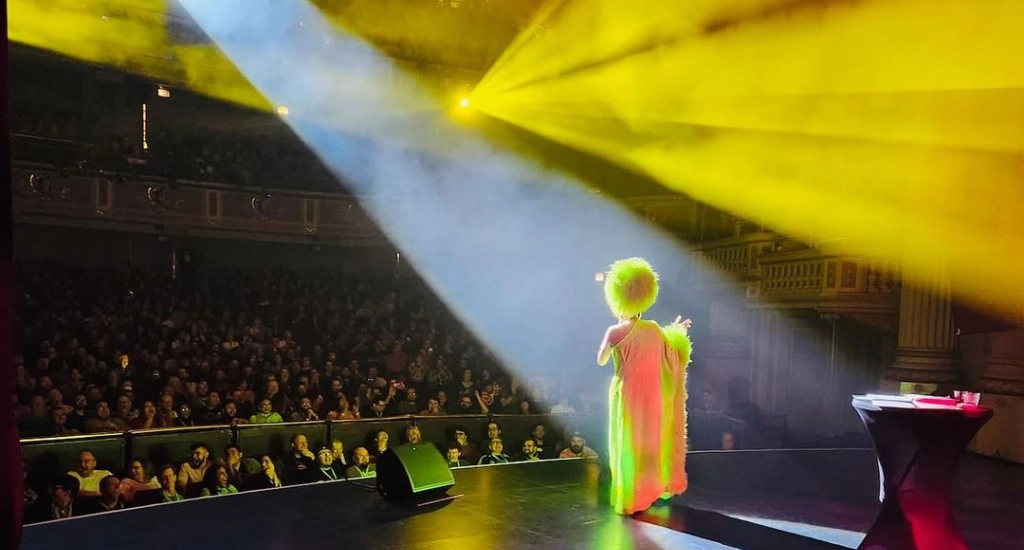 Bianca del Rio iluminada por luzes brancas e amarelas, se apresentando no palco falando para a plateia