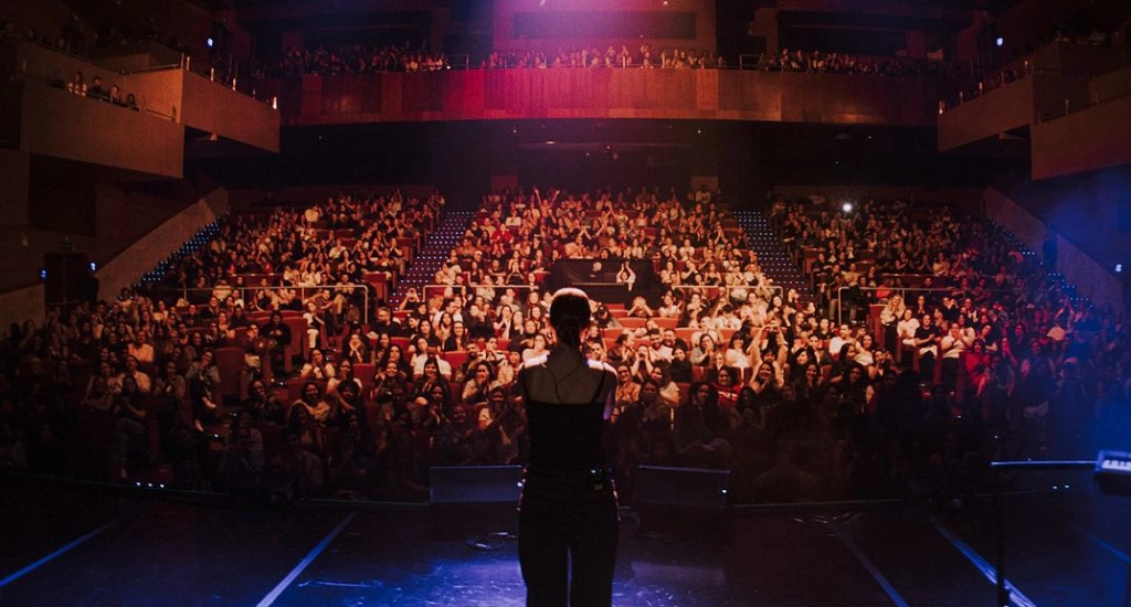 Carol Biazin olhando para o público iluminado com luzes coloridas dentro de um teatro