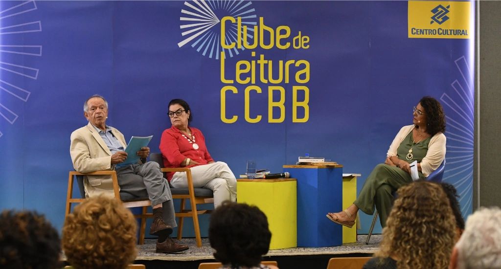 Pessoas conversanso no Clube de Leitura CCBB com mesas, cadeiras e plateia na frente