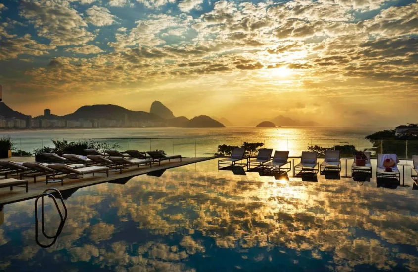 Durante o pôr do sol, área de lazer na cobertura do Fairmont Rio de Janeiro Copacabana com piscina, espreguiçadeiras, vista da praia e céu refletido na piscina