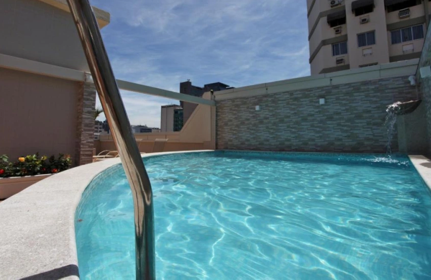 Em um dia de sol, área de lazer do Fluminense Hotel com piscina, cascata e flores decorativas