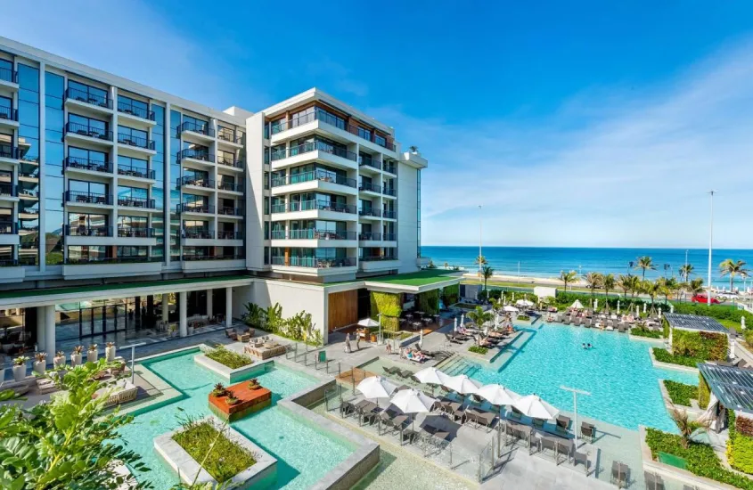 Em um dia ensolarado, vista aérea do Grand Hyatt com piscinas, plantas decorativas, espreguiçadeiras, guarda-sóis e praia na frente