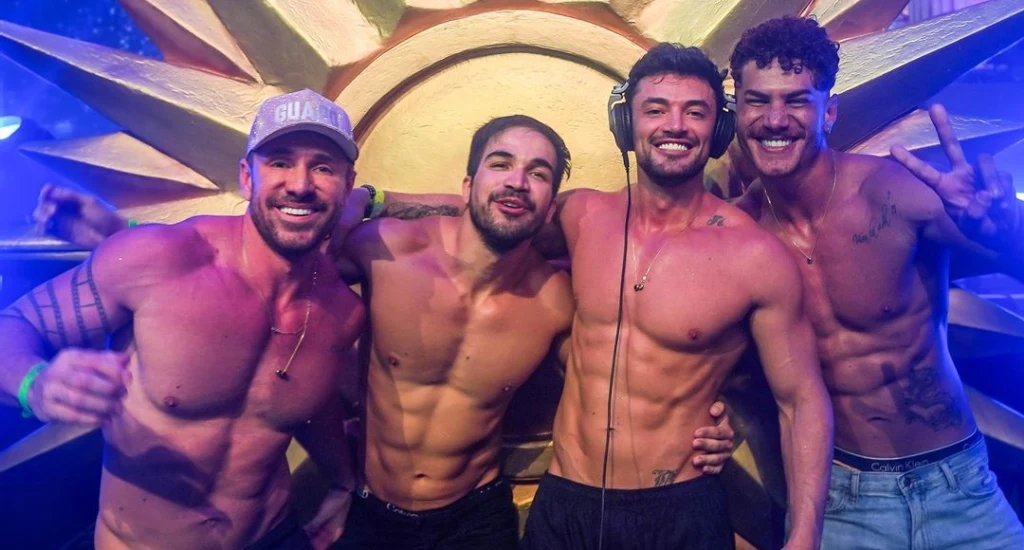 Homens curtindo a festa com luzes coloridas ao redor e mesa de dj na frente