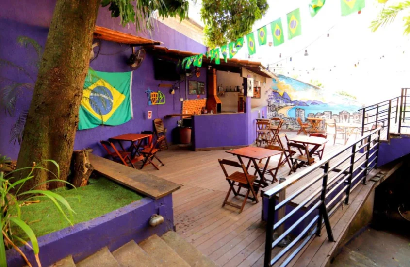 Em um dia de sol, lounge do Pura Vida Hostel com mesas, cadeiras, bandeiras do Brasil, bar e árvores ao redor