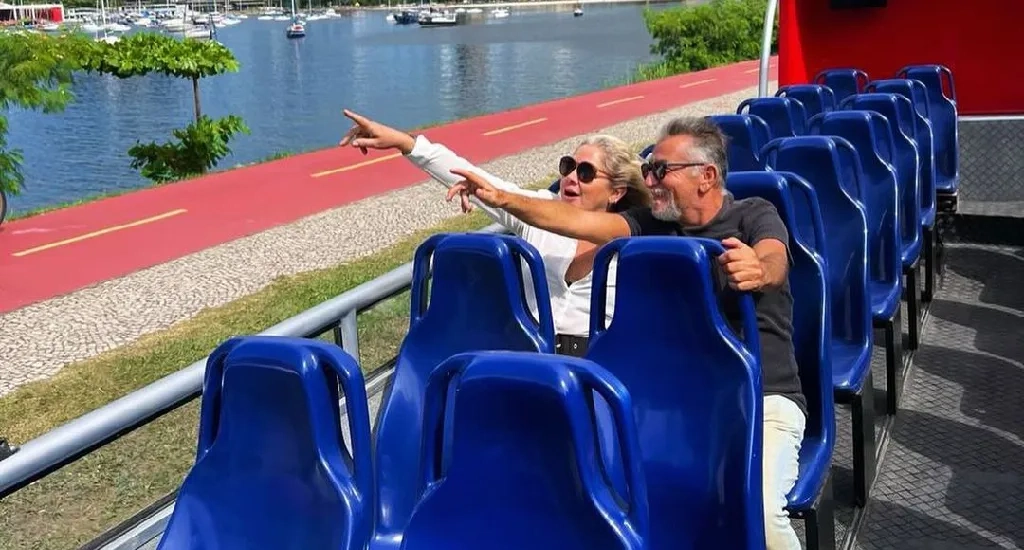 Em um dia de sol,interior do Rio Samba Bus com idosos apontanto para os pontos turísticos da cidade com árvores e mar do lado