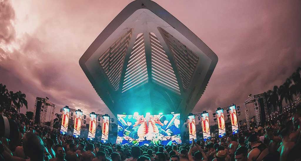 Palco do Rio Ehe New World New Years festival com luzes coloridas, público embaixo, telões de led e árvores ao redor