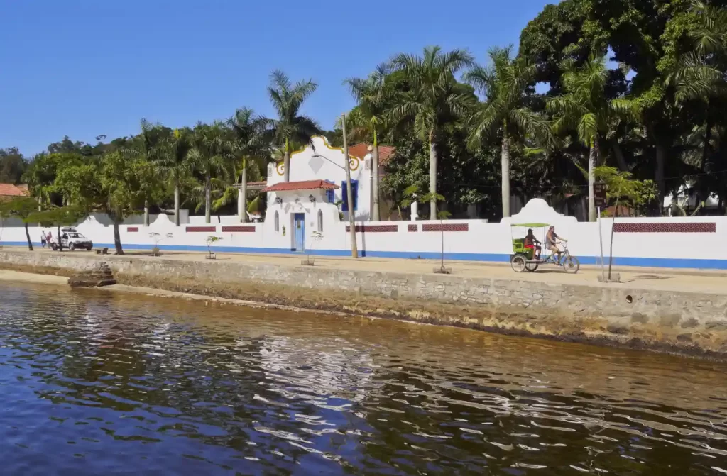 Em um dia de sol, paisagem de Paquetá com lago do lado, pessoas andando de bicicleta e árvores ao redor