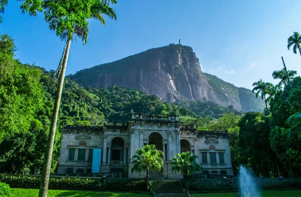 Em um dia ensolarado, fachada do Parque Lage com árvores e flores ao redor, e montanhas no fundo