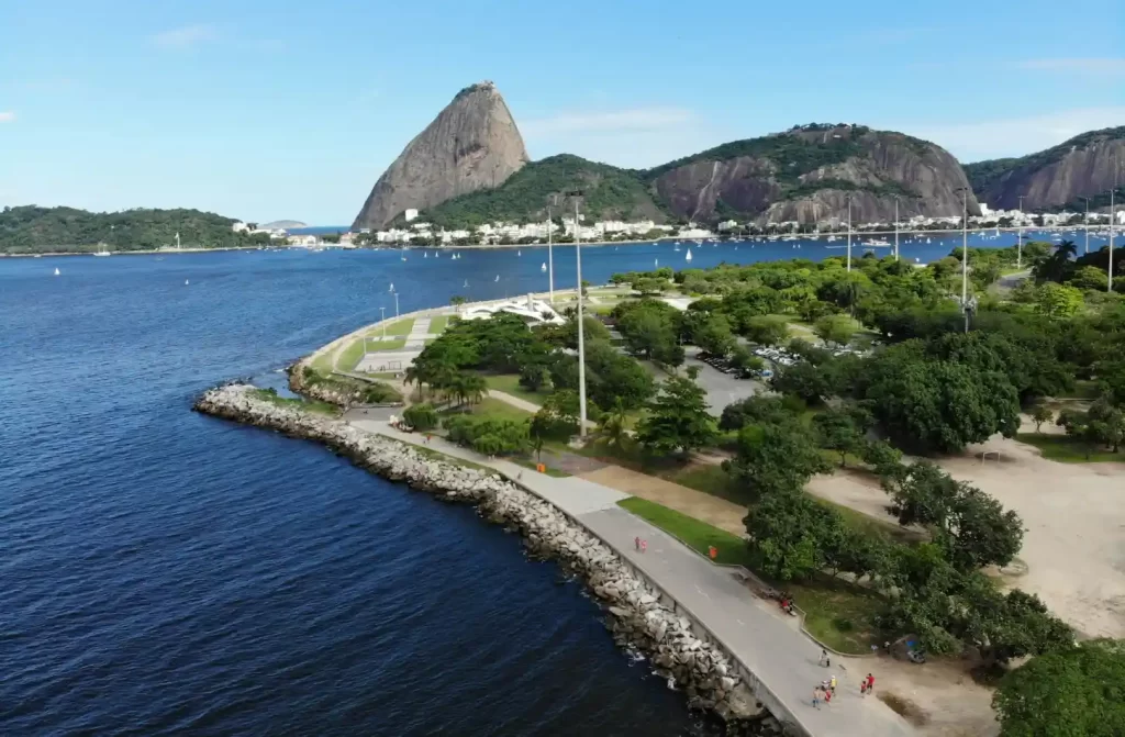 Em um dia ensolarado, vista aérea do aterro do flamengo com mar do lado, montanhas atrás e várias árvores ao redor