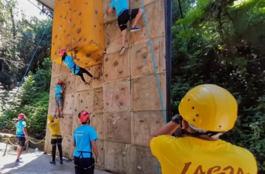 Em um dia de sol, área de escalada do Parque da Catacumba com pessoas subindo, árvores ao redor e instrutores em baixo