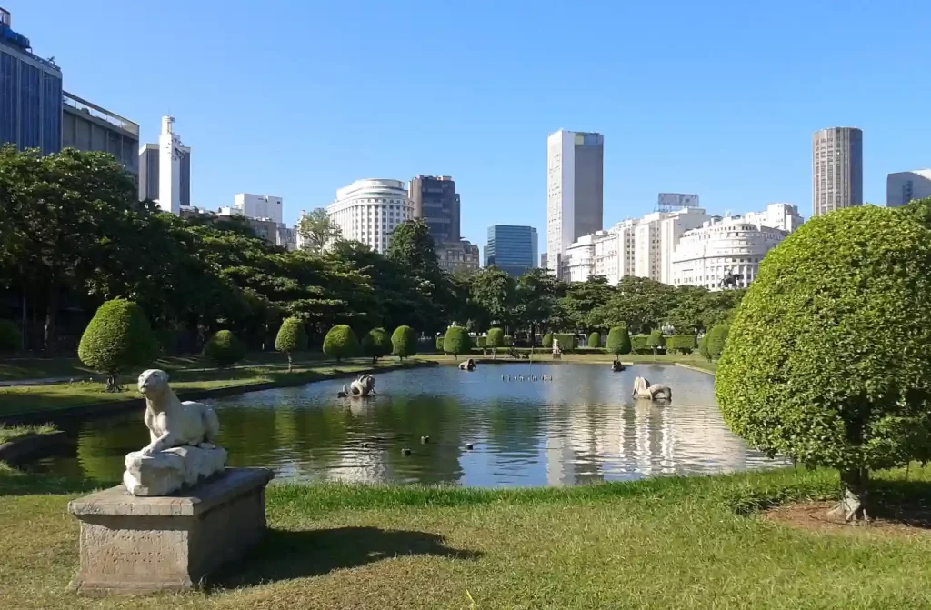 Em um dia ensolarado, lago da Praça Paris com árvores, flores, esculturas e prédios ao redor