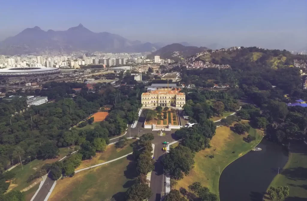 Em um dia de sol, vista aérea da Quinta da Boa Vista com árvores, prédios e lagos ao redor