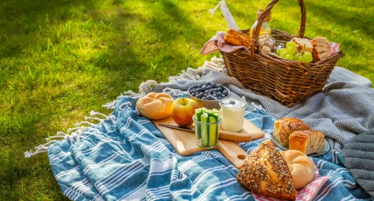 Chão gramado com toalha de picnic, pães, bolos, frutas, cesta, guloseimas e comidinhas