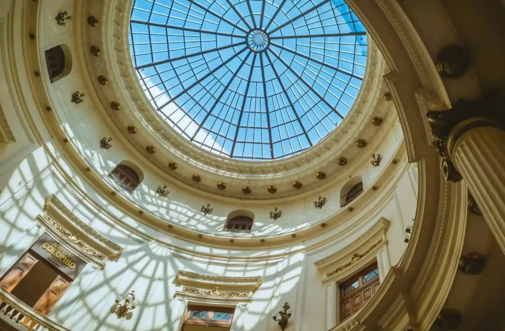 Vista interna do teto de um edifício com uma cúpula de vidro, permitindo a entrada de luz natural, com detalhes arquitetônicos clássicos e sombras projetadas nas paredes.