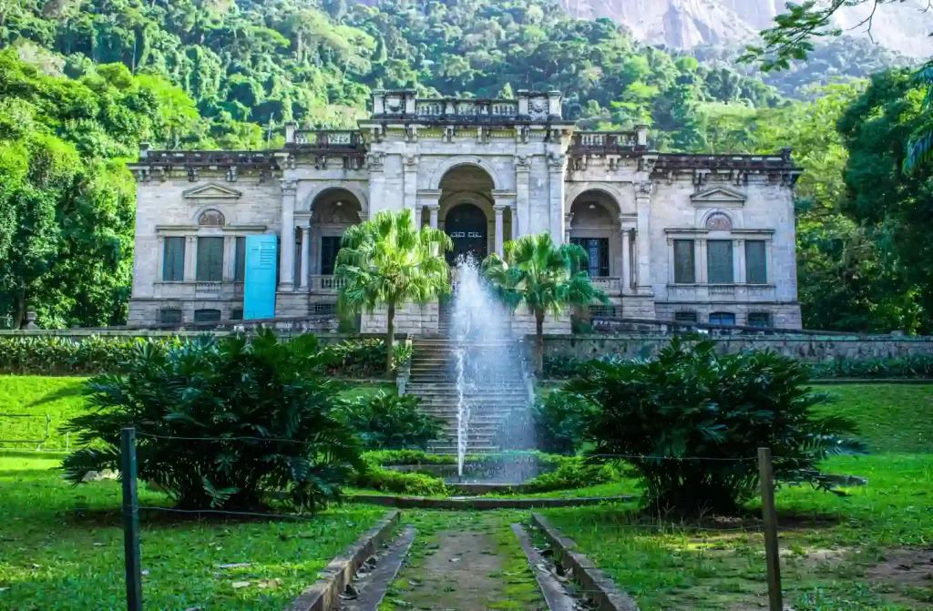 Vista frontal de uma mansão histórica rodeada por árvores, com uma fonte de água no jardim, situada em uma área verde montanhosa.