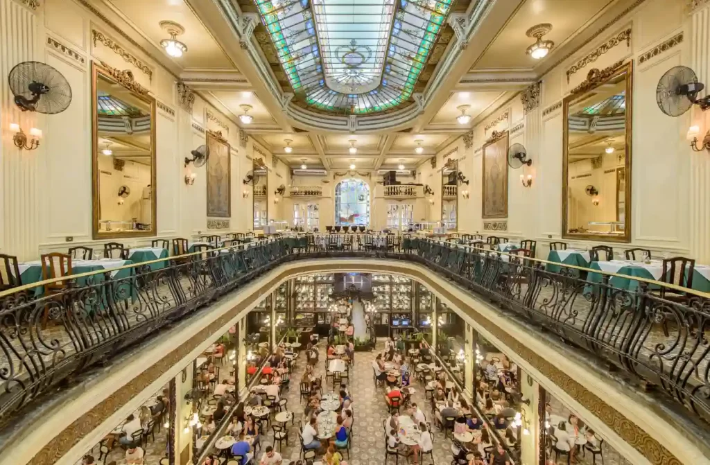 Café histórico de dois andares com arquitetura clássica, grandes espelhos, teto de vidro e varandas ornamentadas, repleto de clientes.