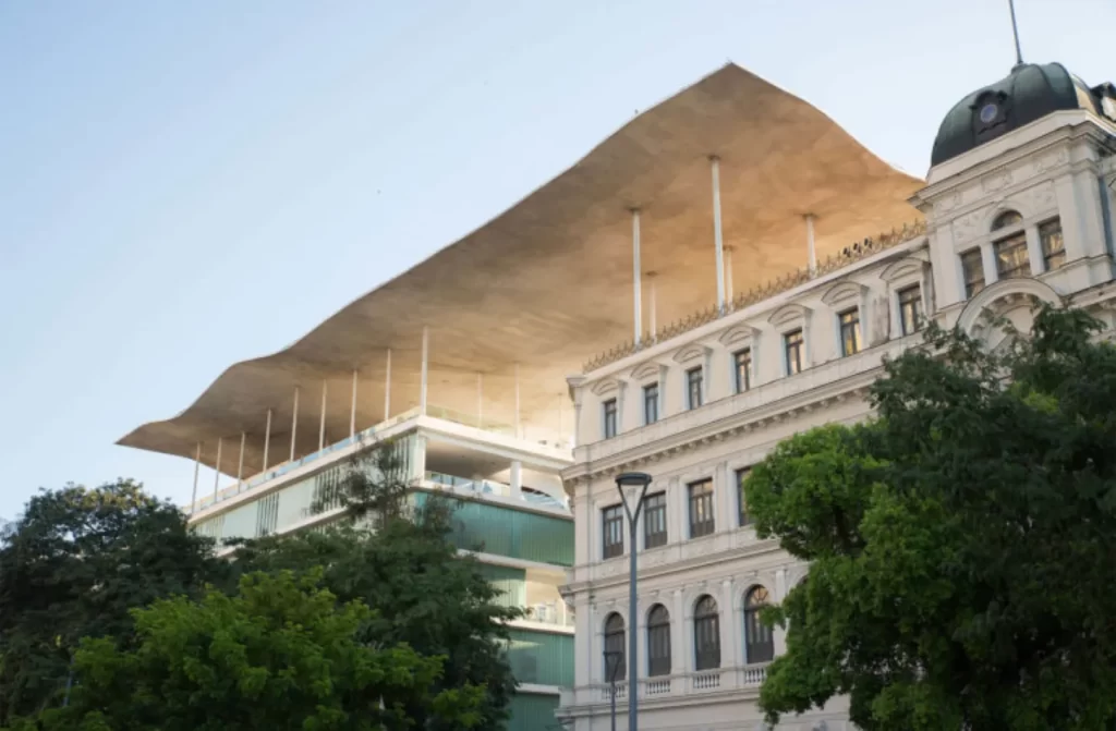 Edifício histórico com fachada clássica ao lado de uma estrutura moderna com grandes vidraças e um amplo teto sustentado por colunas finas.