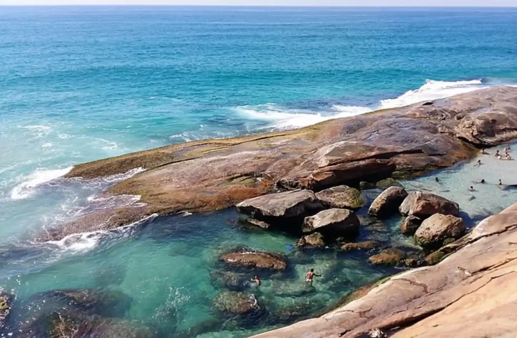 Em um dia ensolarado, praia com pessoas se banhando e pedras ao redor