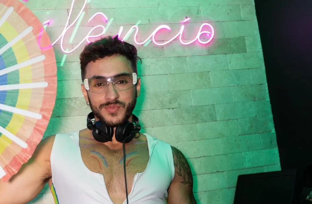 Dj de óculos, fone e leque lgbt tocando no Silêncio Bar, com led atrás