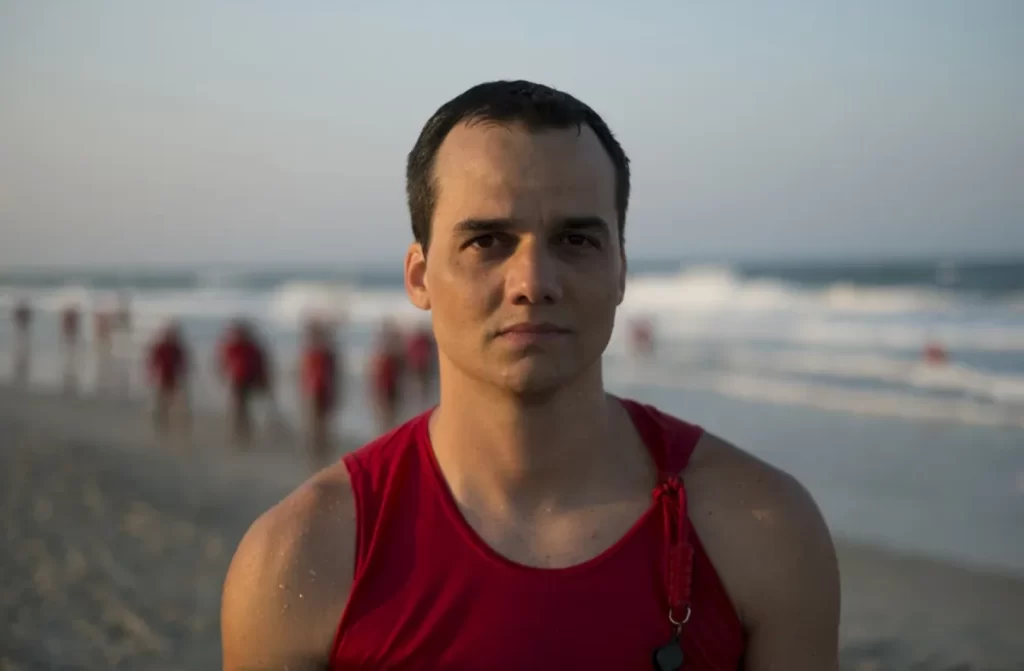 Em um dia de sol, Wagner Moura atuando em a praia do futuro com outros guarda-vidas e praia atrás