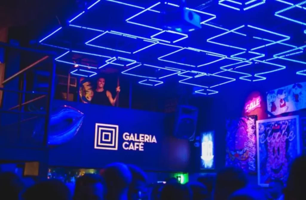 Interior da galeria café com luzes azuis, letreiro da balada e pessoas dentro