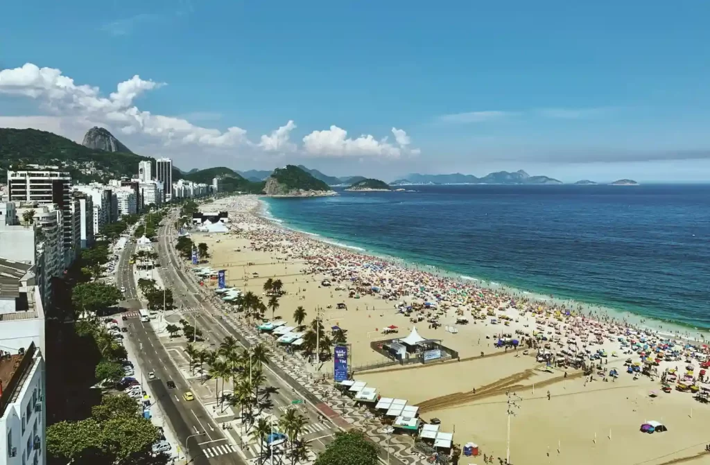 Em um dia ensolarado, paisagem da praia com pessoas, árvores, guarda-sóis, pessoas, carros, tendas e montanhas atrás