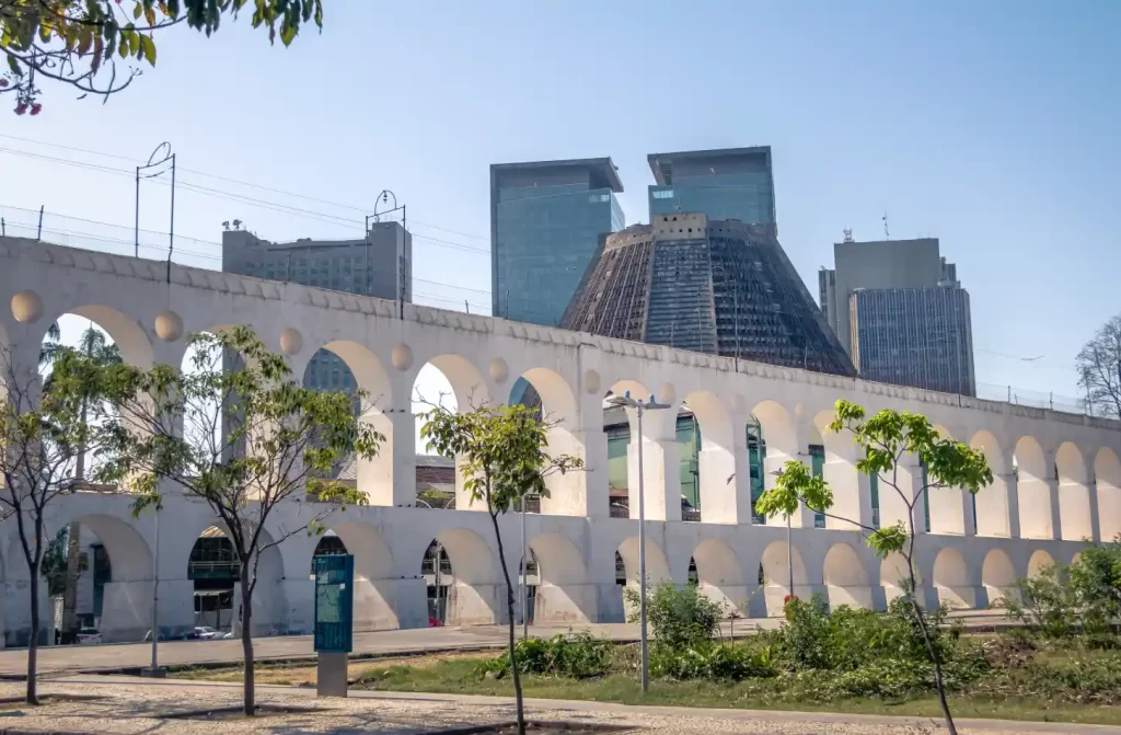 Em um dia de sol, arcos da lapa com árvores ao redor e prédios atrás