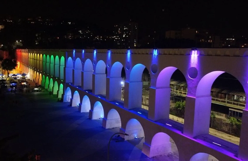 Durante a noite, Arcos da Lapa iluminados com as luzes da bandeira lgbt e árvores ao redor