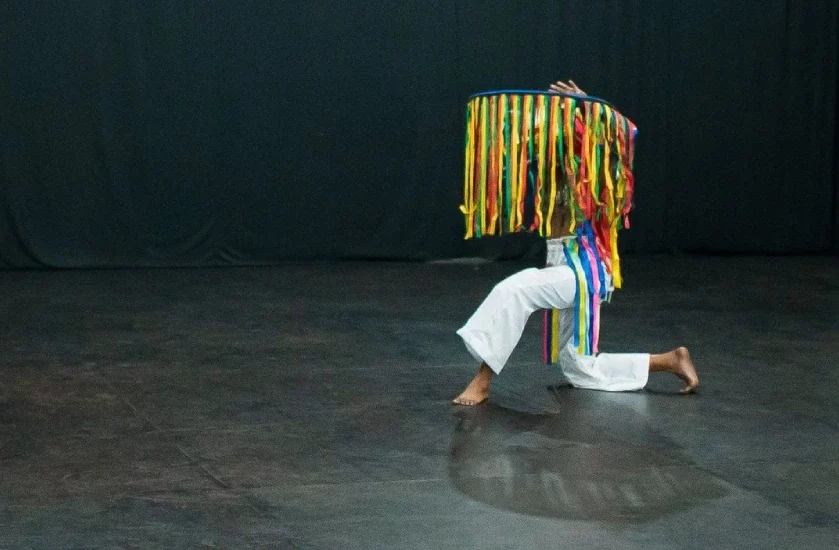 Apresentação no centro das artes com dançarino de calça branca e chapéu com faixas coloridas