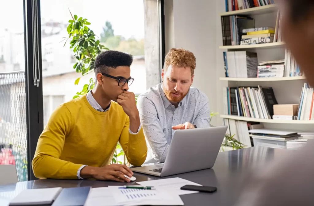 Dois colegas de trabalho discutindo um projeto em um escritório, ambos concentrados em uma tela de laptop, com papéis de relatório e gráficos na mesa.