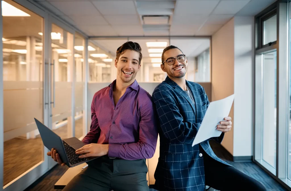 Dois homens sorridentes em um escritório moderno, um segurando um laptop e o outro uma folha de papel, simbolizando um ambiente de trabalho descontraído e colaborativo.