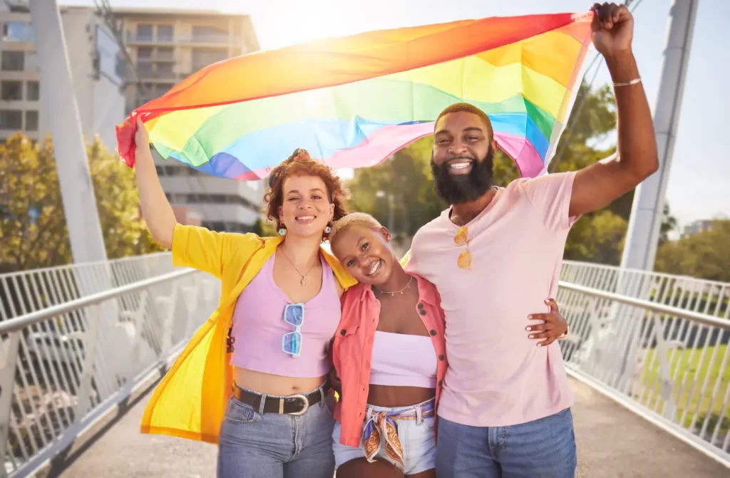 Em um dia ensolarado, 3 pessoas abraçadas segurando a bandeira LGBT com árvores ao redor