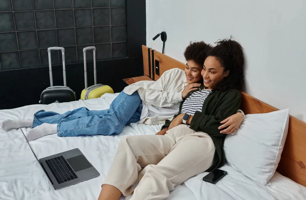 casal lésbico deitado na cama, com malas do lado e computador