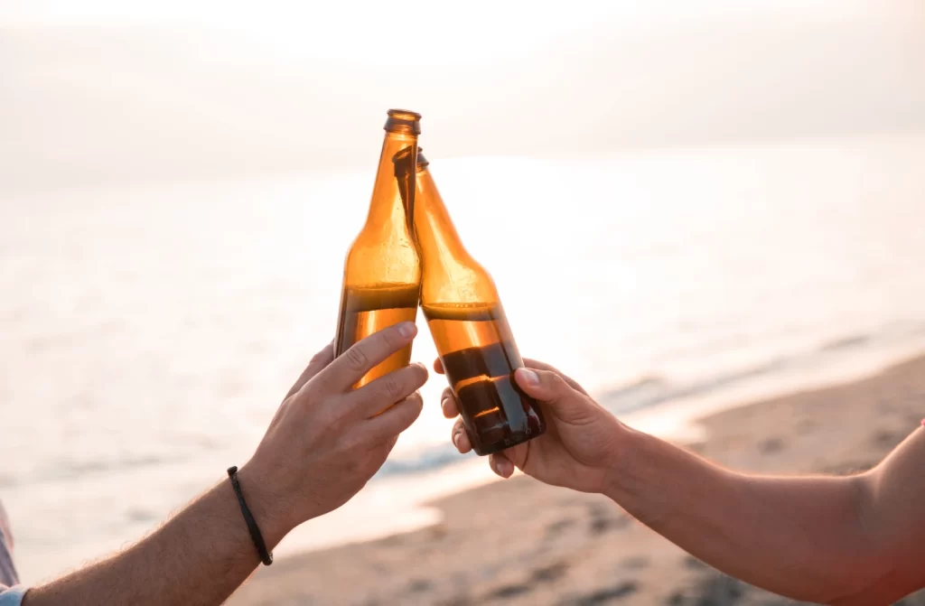 Em um dia ensolarado, pessoas brindando com cervejas e praia no fundo