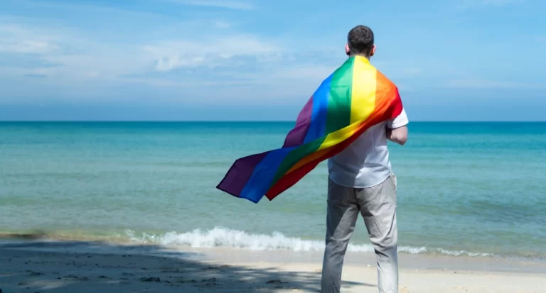 Em um dia ensolarado, pessoa na praia segurando a bandeira LGBT e olhando pro mar