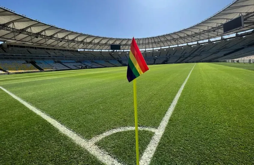 Em um dia ensolarado, gramado do Maracanã  com bandeira lgbt no canto do campo