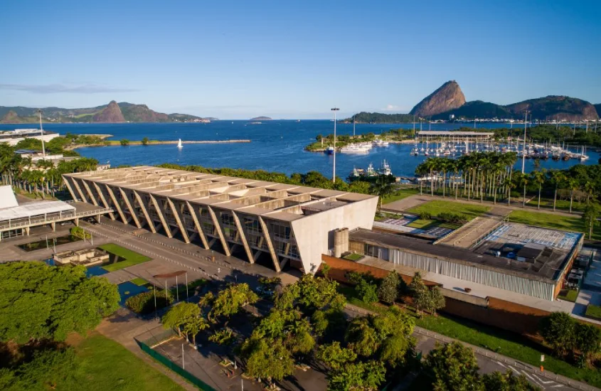 Em um dia ensolarado, vista aérea do Museu de Arte Moderna (MAM) com árvores ao redor e mar do lado