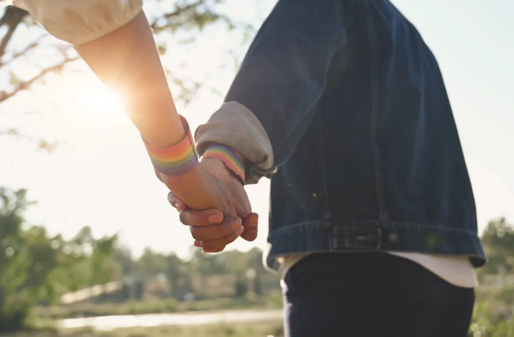 Em um dia de sol, pessoas dando as mãos com árvores ao redor e pulseiras lgbt