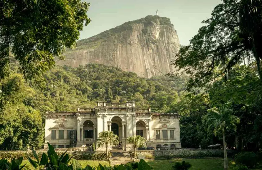 Em um dia de sol, fachada do Parque Lage com flores, árvores e montanhas ao redor