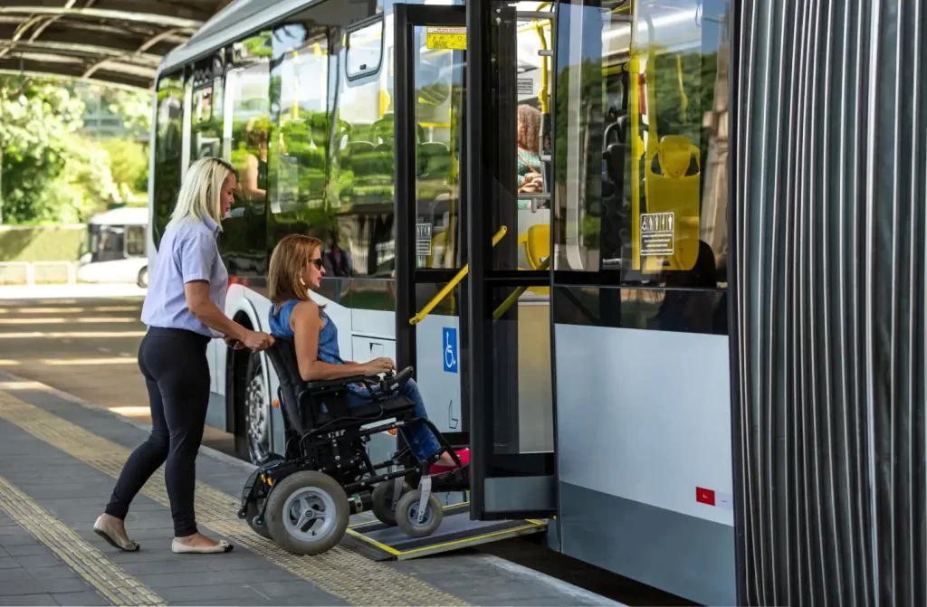 Durante um dia de sol, pessoa ajudando cadeirante a entrar dentro do ônibus com árvores ao redor