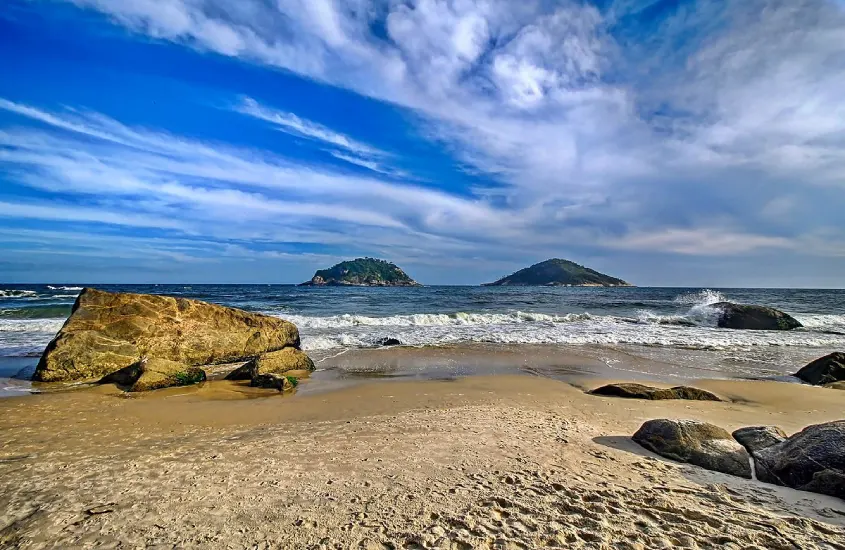 Em um dia ensolarado, Praia do Abricó com pedras, mar quebrando e ilhas atrás