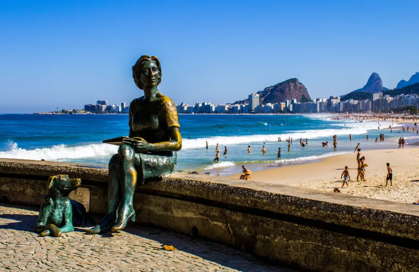 Em um dia de sol, praia do Leme com estátuas famosa, pessoas se banhando e montanhas no fundo