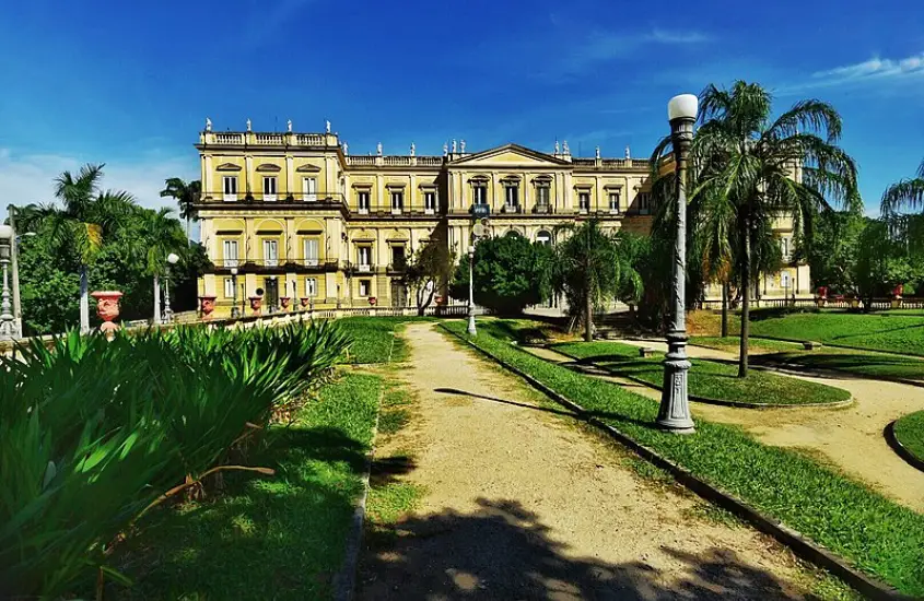 Em um dia de sol, fachada da Quinta da Boa Vista com área gramada, luzes e árvores ao redor