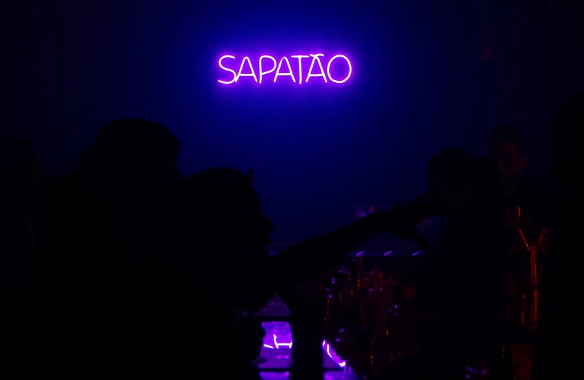 Letreiro de led colorido escrito "sapatão" e duas pessoas no Tau Bar Club