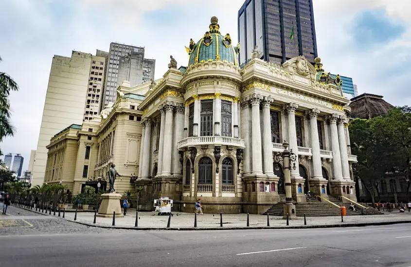 Em um dia de sol com nuvens, fachada do Teatro Municipal com pessoas e árvores ao redor