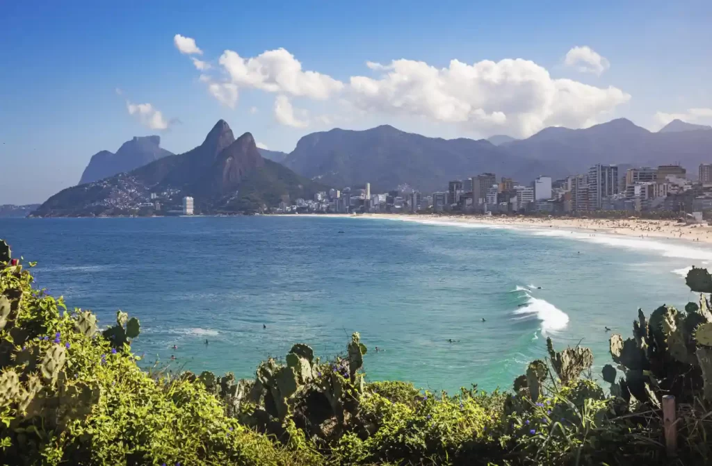 Em um dia de sol com nuvens, paisagem da praia com montranhas atrás e árvores ao redor