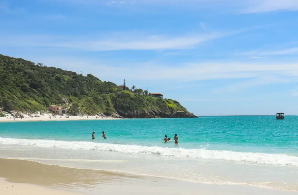 Em uma manhã ensolarada, praia com águas cristalinas, pessoas se banhando e árvores atrás