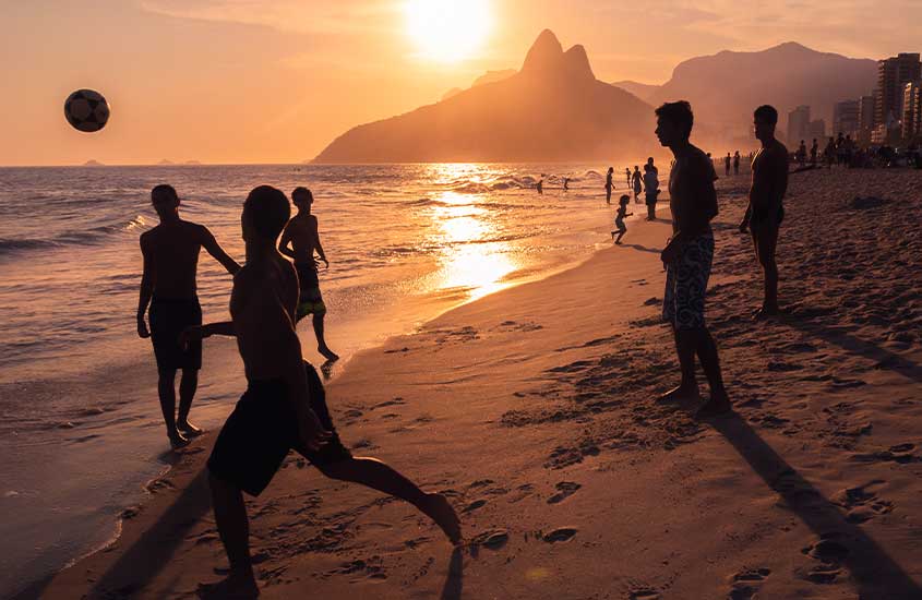 Durante o pôr do sol, pessoas curtindo a praia e jogando bola com montanhas no fundo