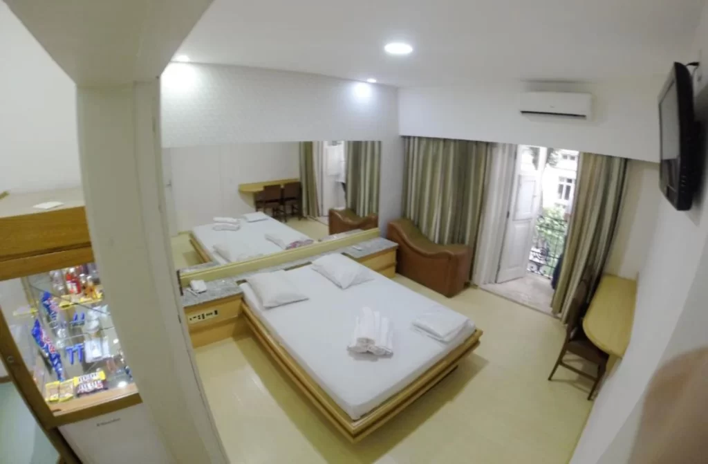 Quarto do motel com cama de casal, cristaleira, TV, cortinas e móveis de madeira